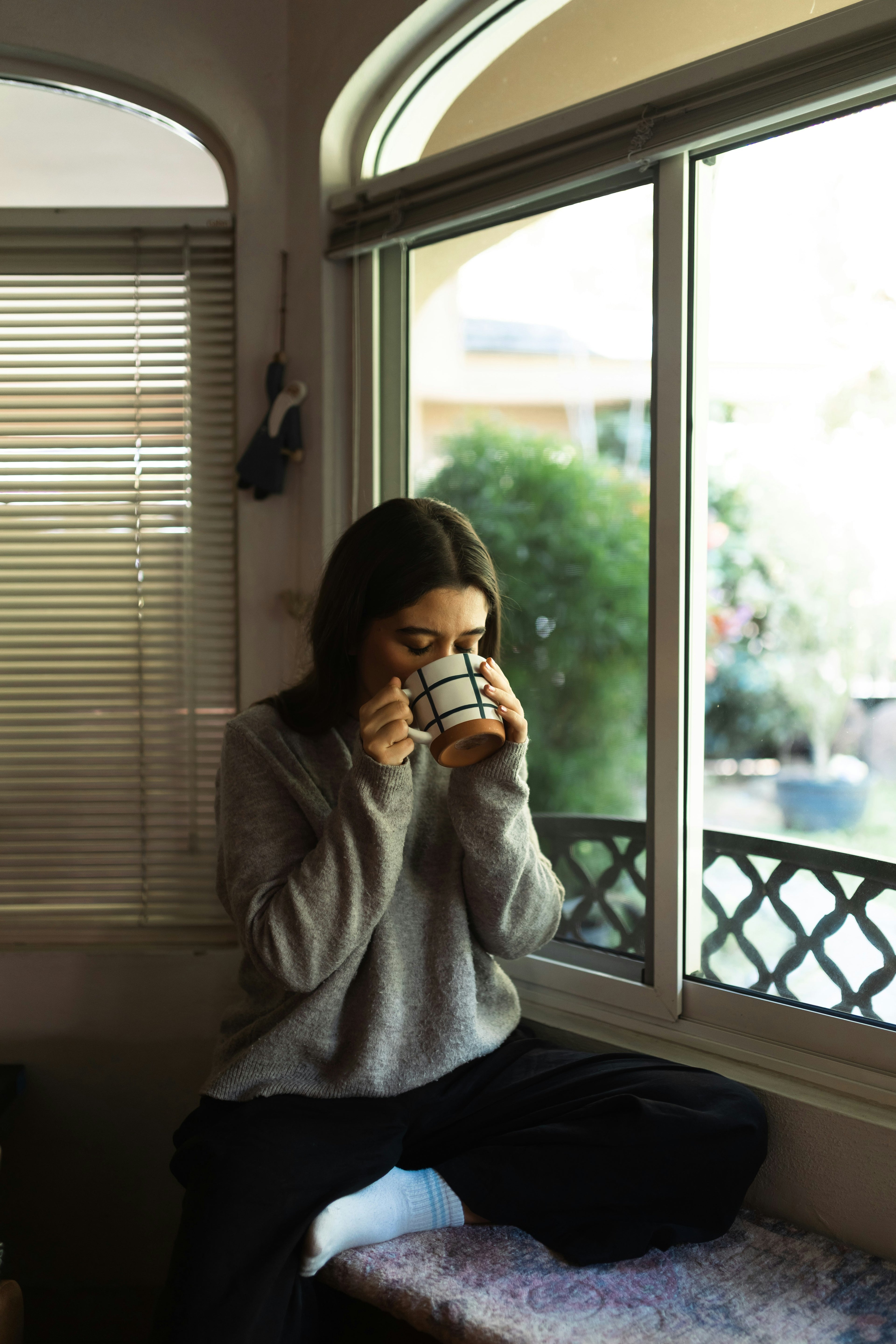 Person sitting by a window holding a mug, wearing a gray sweater and dark pants.