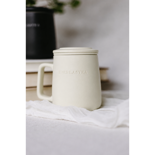 A matte cream-colored ceramic mug with a matching lid and handle, featuring the embossed brand name 'EVERLASTEA' on the front. The mug is styled on a white textured surface with a piece of white fabric and a blurred black pot in the background.