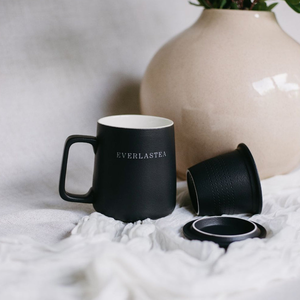A matte black ceramic mug with a handle, featuring the brand name 'EVERLASTEA' printed in white on the side. The mug is shown with its matching black lid and inner cup beside it on a white textured fabric surface, with a beige ceramic vase and green leaves in the background.