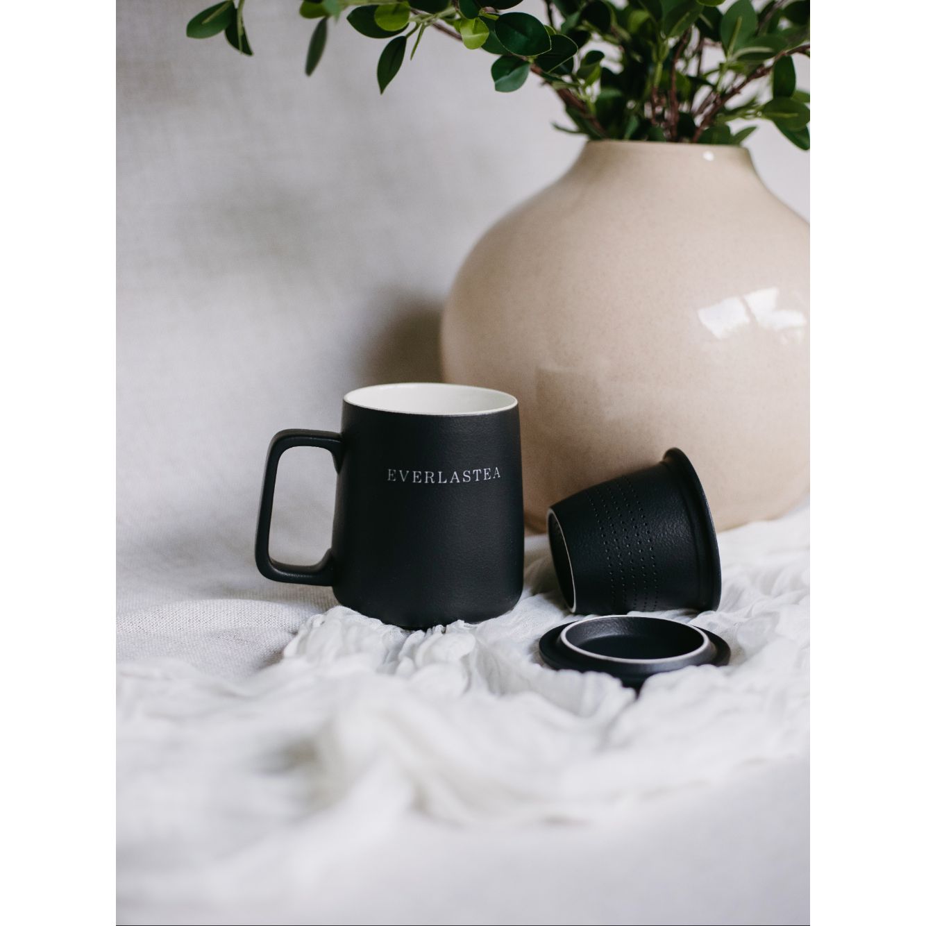 A matte black ceramic mug with a handle, featuring the brand name 'EVERLASTER' printed in white on the side. The mug is shown with its matching black lid and inner cup beside it on a white textured fabric surface, with a beige ceramic vase and green leaves in the background.