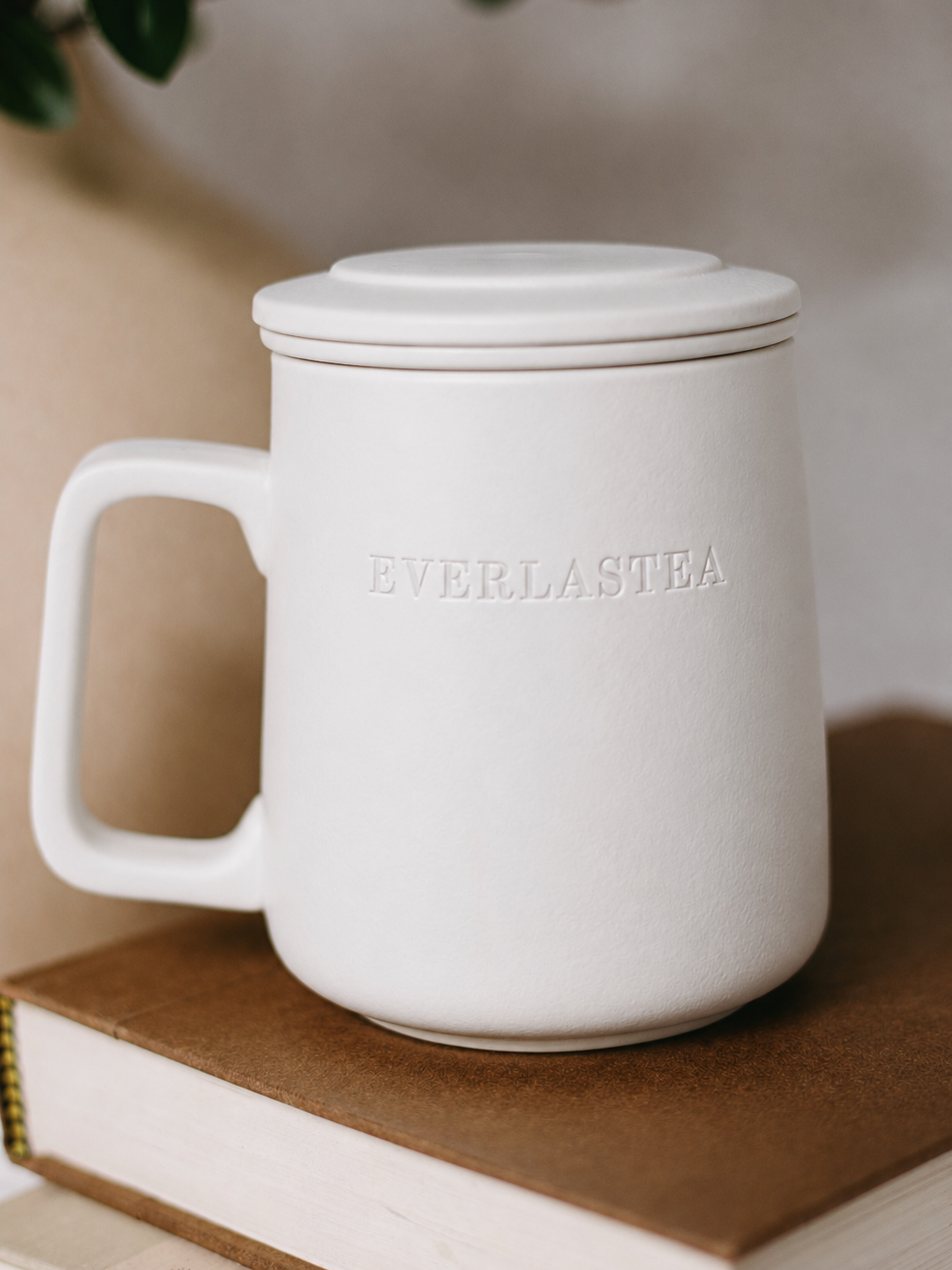 A matte white ceramic mug with a matching lid, featuring the brand name 'EVERLASTEA' embossed on the front, resting on a brown book.
