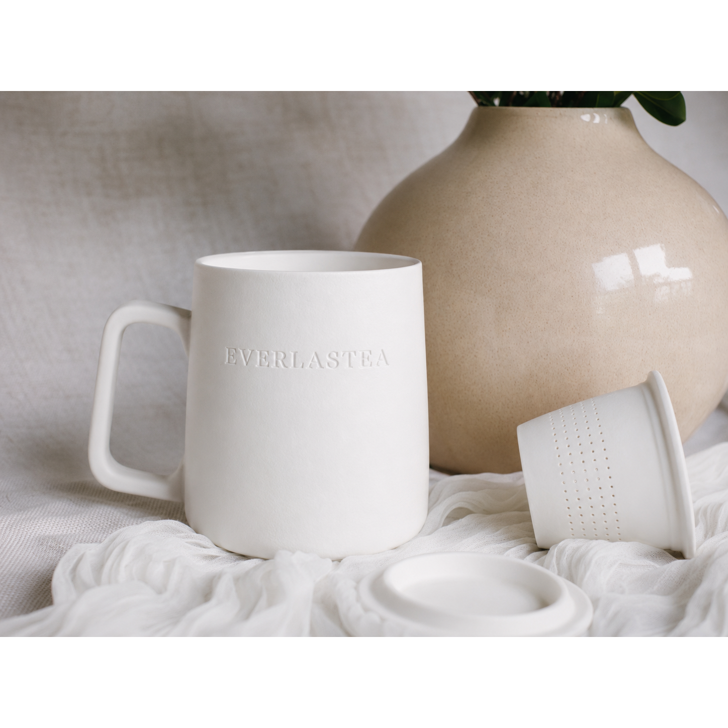 A matte white ceramic mug with the brand name 'EVERLASTEA' embossed on the front, shown alongside its matching lid and infuser insert on a draped white fabric surface, with a beige ceramic vase in the background.