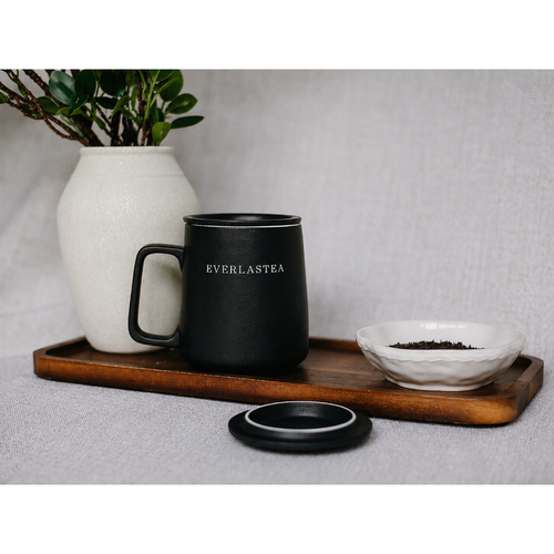 A matte black ceramic travel mug with the brand name 'EVERLASTEA' printed in white on the front, resting on a wooden tray next to a white ceramic bowl of loose tea and a white vase with green leaves, all set against a neutral gray fabric background.