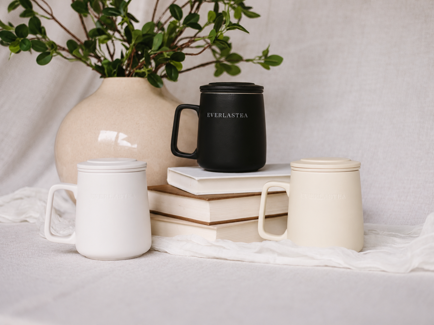 Three ceramic mugs with lids are arranged on a draped white fabric surface with stacked books and a vase of greenery in the background. One mug is white, one is black with the word EVERLASTEA printed on it, and one is warm cream.