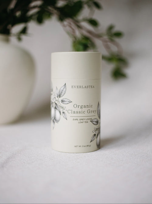A white cylindrical tin of Everlastea Organic Classic Grey leaf tea with black botanical illustrations, set on a white textured surface with a blurred white vase and green leaves in the background.