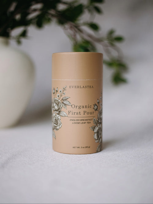 A beige cylindrical tea tin labeled 'EVERLASTEAA Organic First Pour' with floral illustrations, standing on a light surface with a softly blurred white vase and greenery in the background.