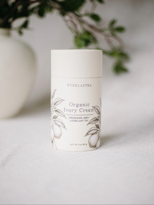 A white cylindrical container of Everlastea Organic Ivory Cream, featuring delicate botanical illustrations of leaves and flowers, set against a soft, blurred background with green foliage.