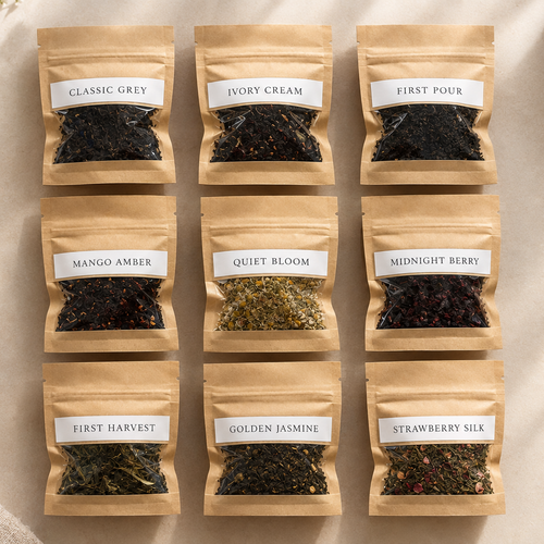Nine small kraft paper tea bags arranged in a 3-by-3 grid on a light fabric surface, each labeled with a different tea variety: Classic Grey, Ivory Cream, First Four, Mango Amber, Quiet Bloom, Midnight Berry, First Harvest, Golden Jasmine, and Strawberry Silk. A cup of tea, flowers, and a bowl of loose tea are placed around the bags as styled props.